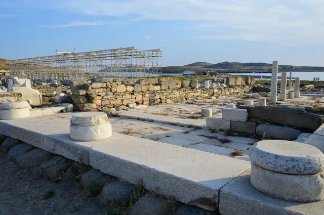 Stone walls, paving and column remains of the Oikos of the Naxians on Delos, with the sea behind.
