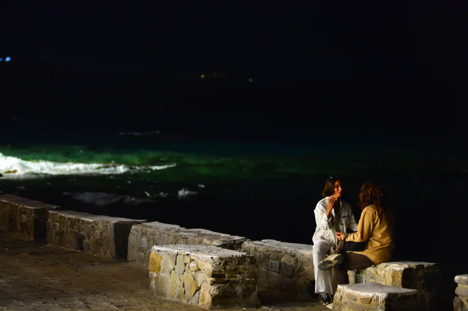 Δύο άνθρωποι κάθονται σε πέτρινο στηθαίο δίπλα στη σκοτεινή θάλασσα στην προκυμαία της Χώρας