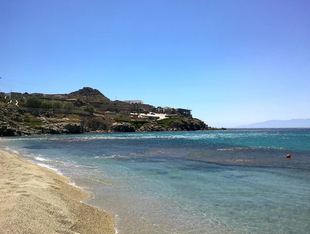 Sand and turquoise water beneath a rocky promontory with buildings at Paradise Beach